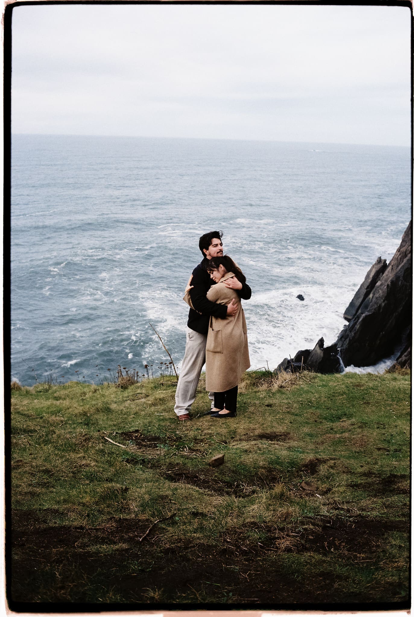 couple shares a hug at the edge of a west coast cliff with the ocean stretching out through the background
