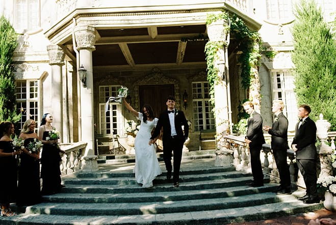 bride and groom exit their wedding venue as their guests celebrate